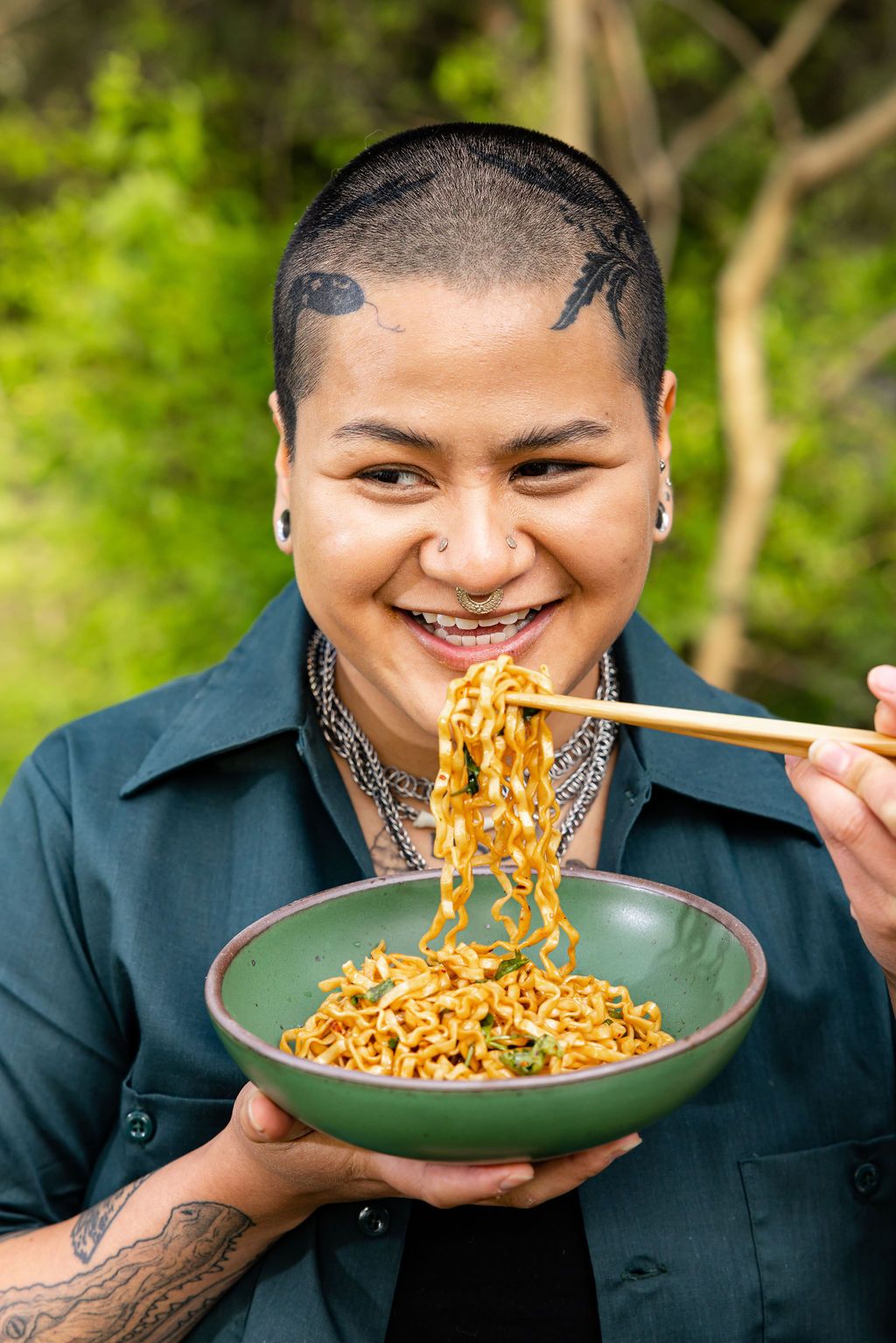 A person with a shaved head is smiling and holding a bowl of noodles and a pair of chopsticks