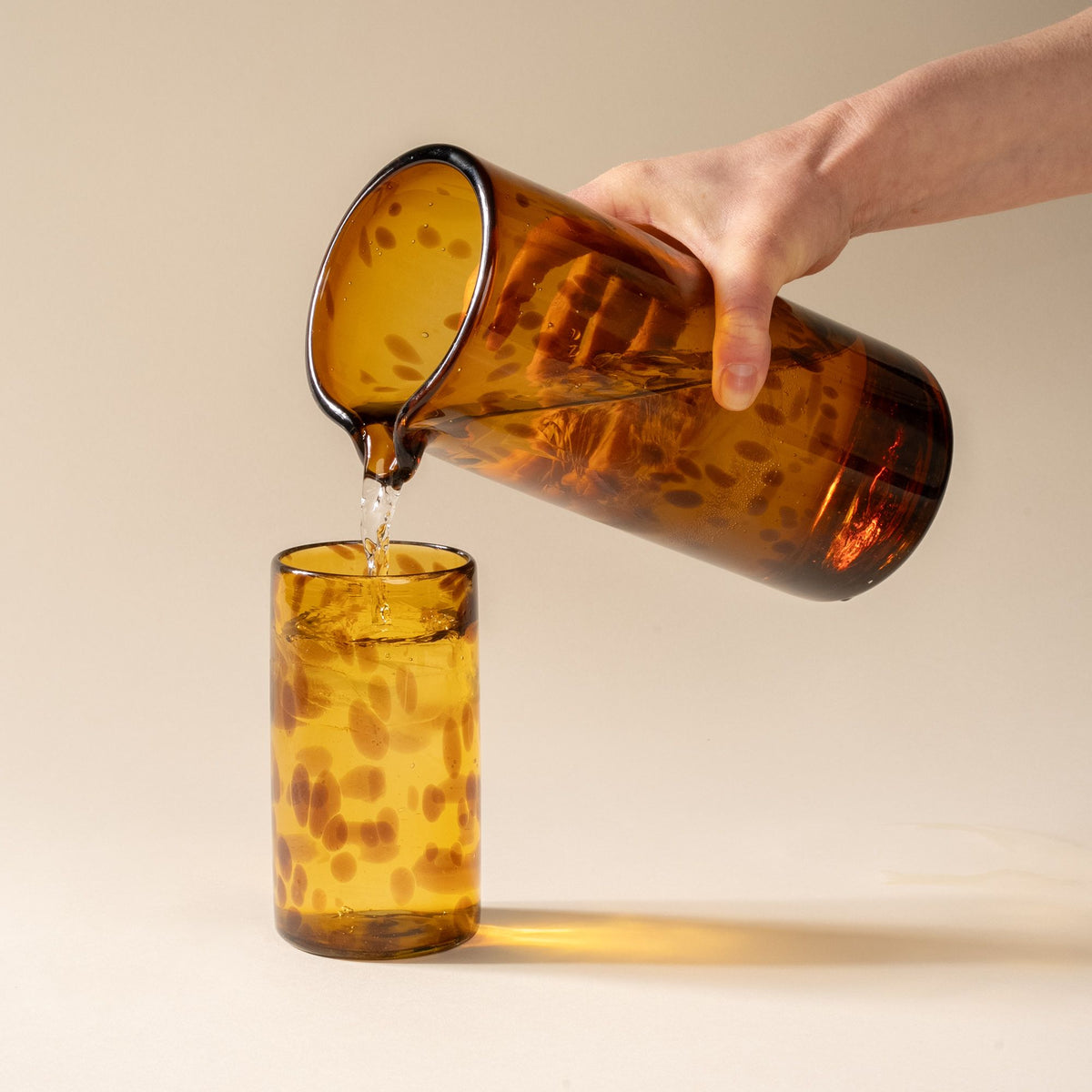 A hand holds a tortoiseshell transparent glass pitcher with small spout at the top, and pours a drink into a tall tumbler.