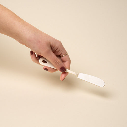 A hand holding a butter knife made of white enamel.