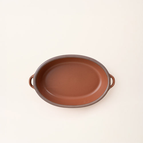 A medium, oval, matte, terracotta ceramic baking dish with two side handles, on a solid off-white background, photographed from overhead. 