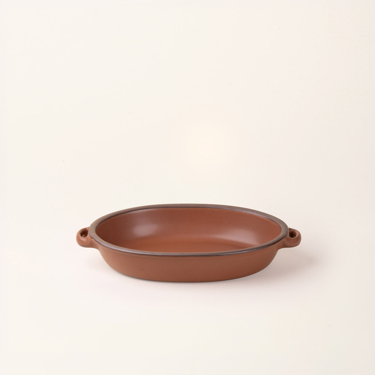 A medium, oval, matte, terracotta ceramic baking dish with two side handles, on a solid off-white background.