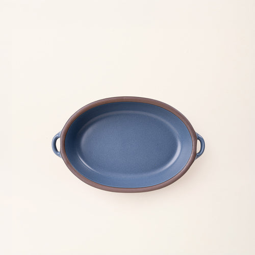 A medium, oval, matte, grounded blue ceramic baking dish with two side handles, on a solid off-white background, photographed from overhead. 