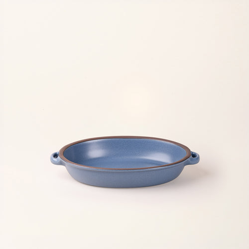 A medium, oval, matte, grounded blue ceramic baking dish with two side handles, on a solid off-white background.