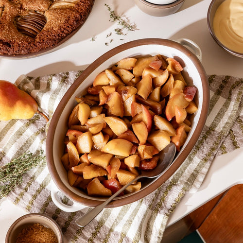 An overhead view of a medium oval, cool white, ceramic baking dish with handles, filled with cooked sliced apples and a serving spoon, resting on a striped kitchen towel surrounded by baked goods, fresh apples, and herbs.