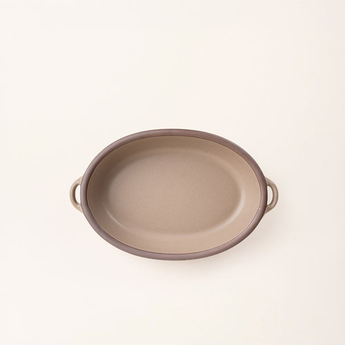A medium, oval, matte, warm pale brown ceramic baking dish with two side handles, on a solid off-white background, photographed from overhead. 