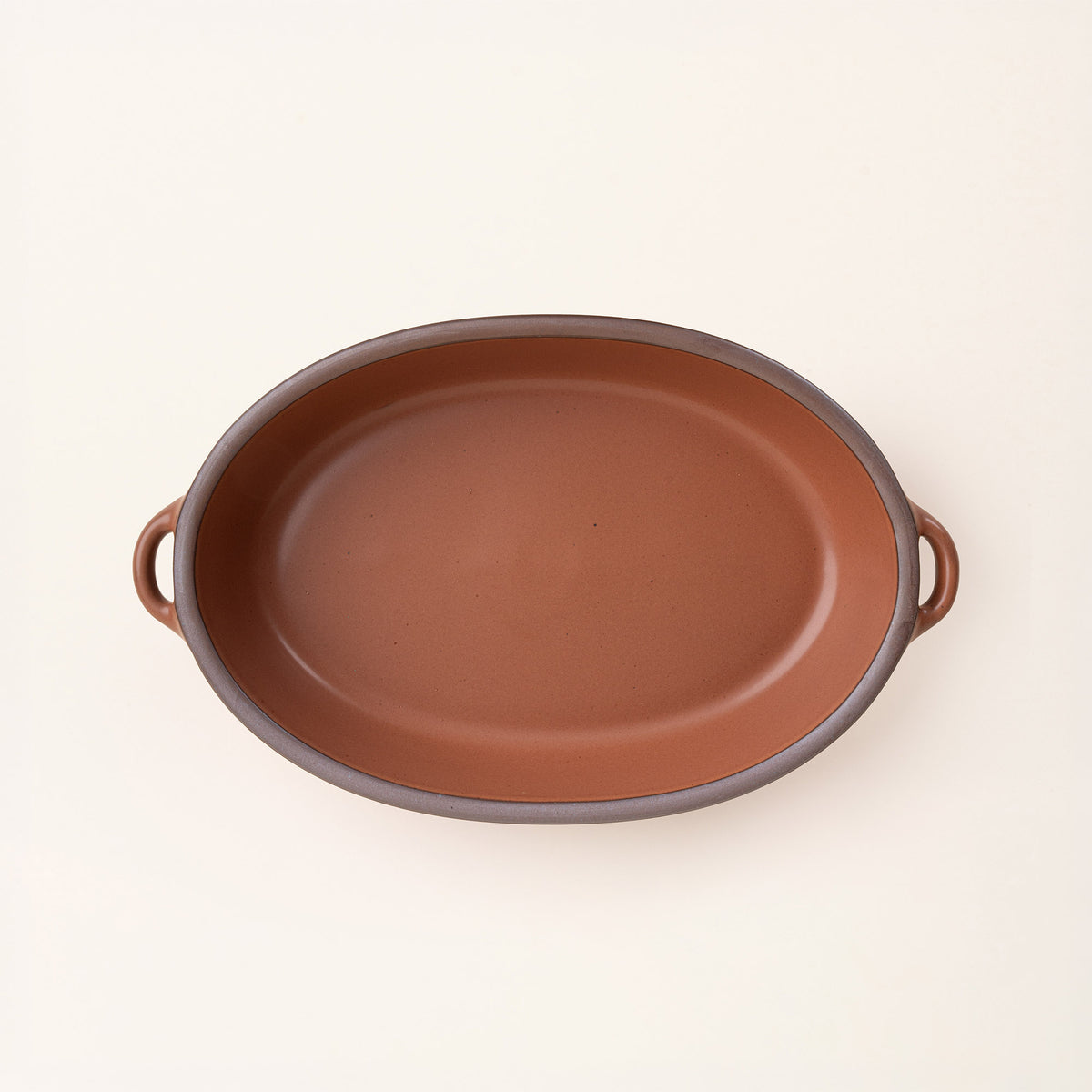 A large, oval, matte, cool terracotta ceramic baking dish with two side handles, on a clean, solid off-white background.