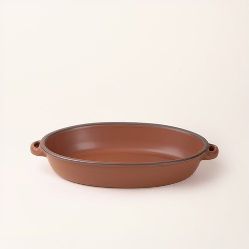 A large, oval, matte, terracotta ceramic baking dish with two side handles, on a solid off-white background.