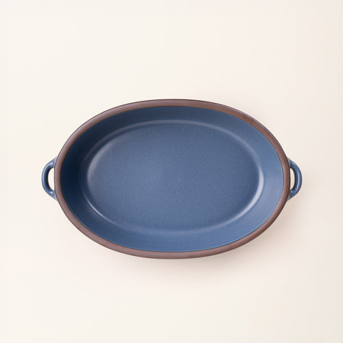 A large, oval, matte, grounded blue ceramic baking dish with two side handles, on a solid off-white background, photographed from overhead. 