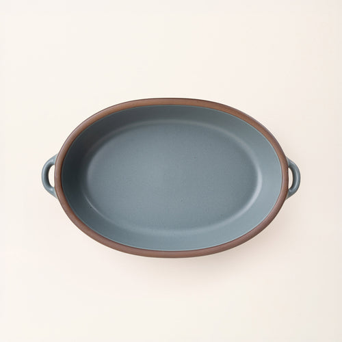 A large, oval, matte, blue-grey ceramic baking dish with two side handles, on a solid off-white background, photographed from overhead. 