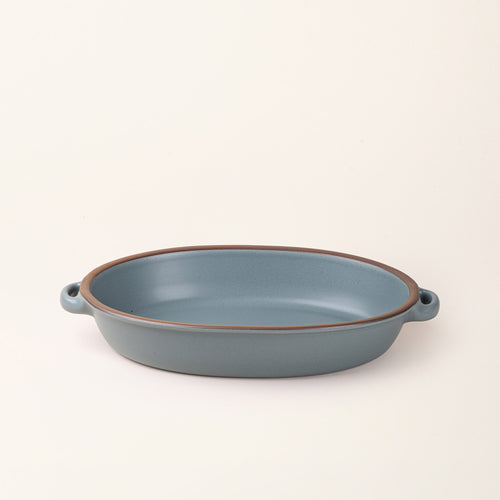 A large, oval, matte, blue-grey ceramic baking dish with two side handles, on a solid off-white background, photographed from overhead. 