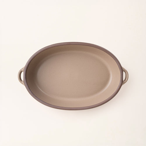 A large, oval, matte, warm pale brown ceramic baking dish with two side handles, on a clean, solid off-white background.