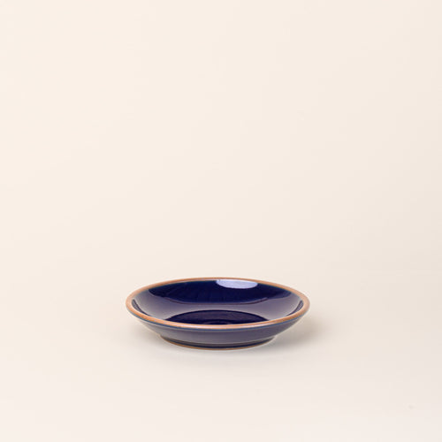 A small ceramic plate with a curved bowl edge in a glossy dark blue color featuring iron speckles and an unglazed rim.