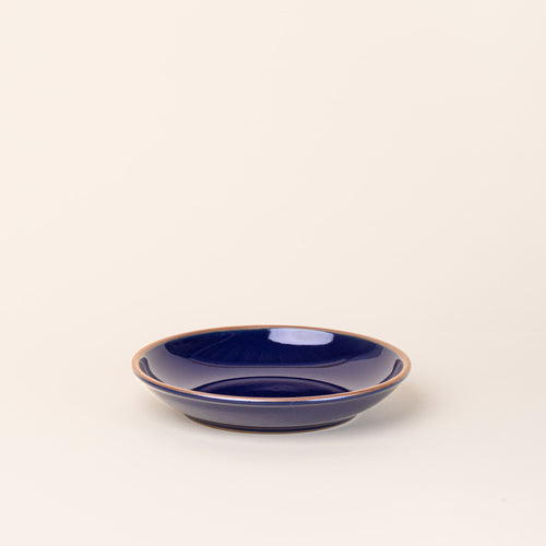 A medium ceramic plate with a curved bowl edge in a glossy dark blue color featuring iron speckles and an unglazed rim.