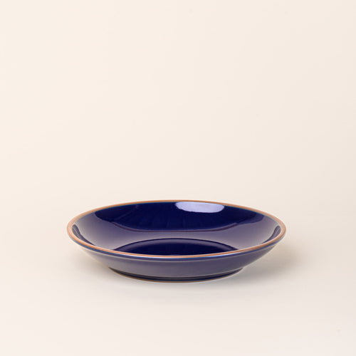 A large ceramic plate with a curved bowl edge in a glossy dark blue color featuring iron speckles and an unglazed rim.