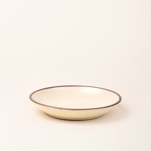 A large ceramic plate with a curved bowl edge in a cream color featuring iron speckles and an unglazed rim.
