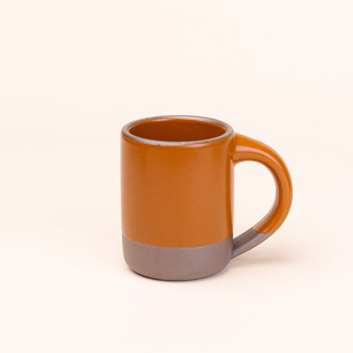 A medium sized ceramic mug with handle in an earthy orange color featuring iron speckles and unglazed rim and bottom base.