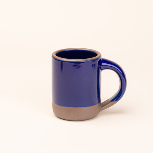 A medium sized ceramic mug with handle in a glossy dark blue color featuring iron speckles and unglazed rim and bottom base.