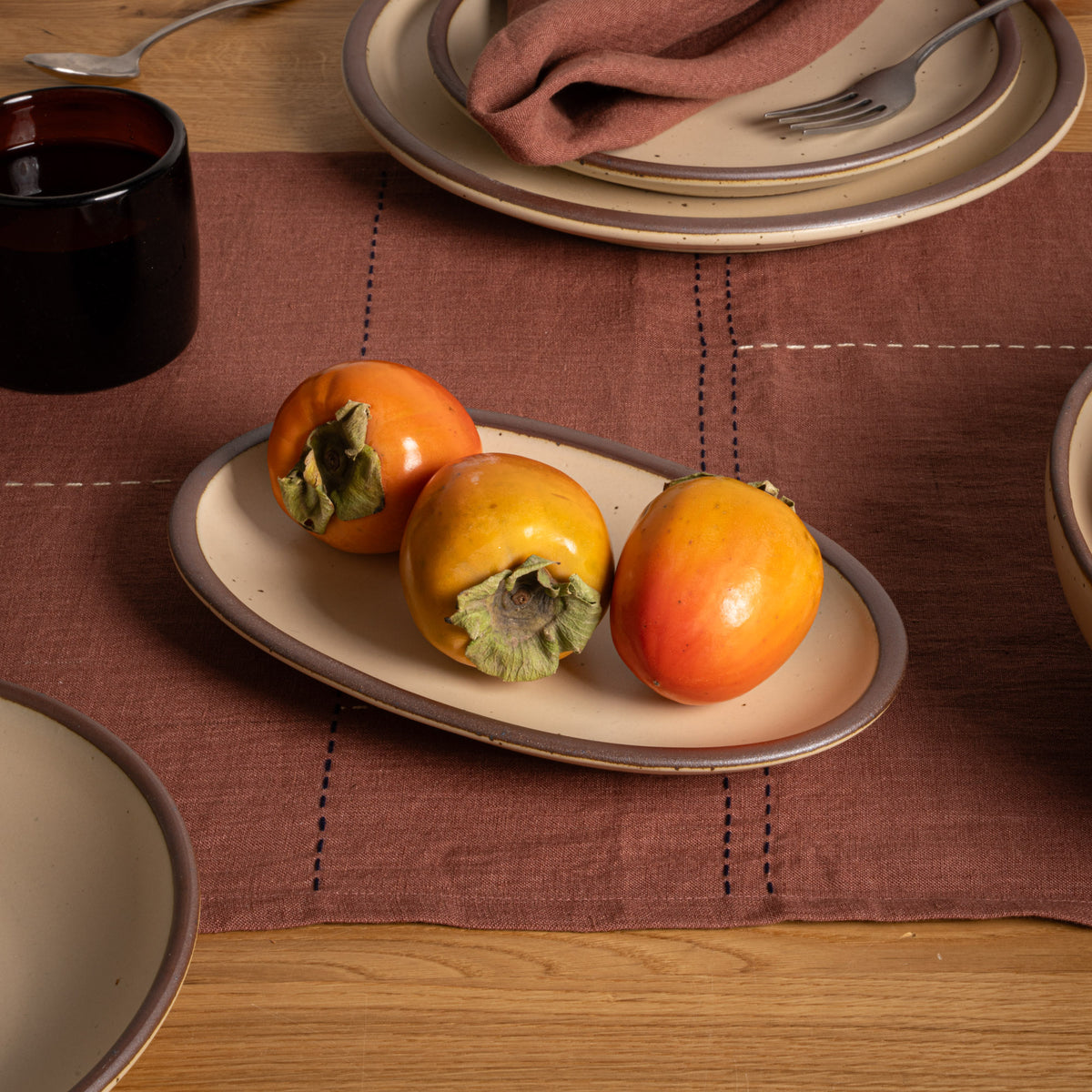 A small oval cream colored ceramic serving platter with an unglazed rim, styled on a brown linen placemat with three persimmon fruits, set on a wooden dining table alongside other plates and a dark glass cup.