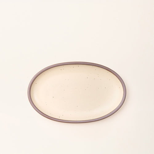 A medium oval ceramic platter in a cream color featuring iron speckles and an unglazed rim