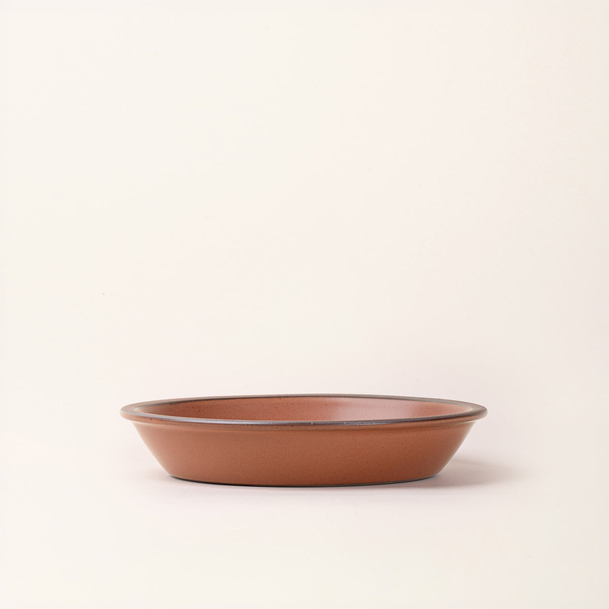 A side view of a shallow round ceramic pie plate in a terracotta color featuring an unglazed rim, against a cream background.