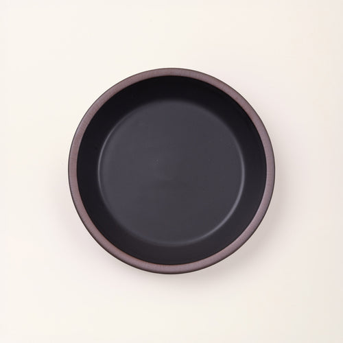 An overhead view of a shallow round ceramic pie plate in a graphite black color featuring an unglazed rim, against a cream background.