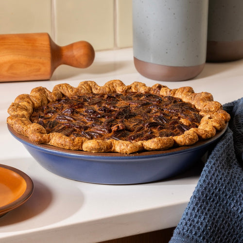 A baked pecan pie in a blue ceramic pie dish on a white countertop, with a wooden rolling pin, speckled mug, and blue cloth in a kitchen setting.