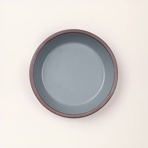 An overhead view of a shallow round ceramic pie plate in a blue-grey color featuring an unglazed rim, against a cream background.