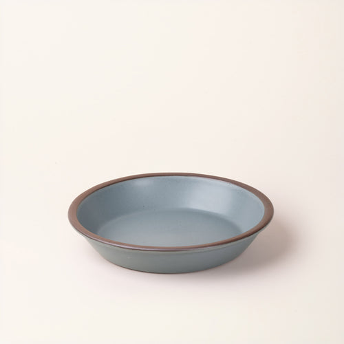 A shallow round ceramic pie plate in a matte blue-grey color featuring an unglazed rim, photographed against a cream background.