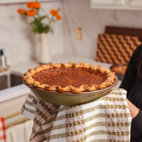 A baked pumpkin pie in an olive green ceramic pie dish held by someone with a stripe kitchen towel in a kitchen.
