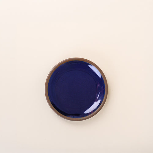 A dessert sized ceramic plate in a glossy dark blue color featuring iron speckles and an unglazed rim.