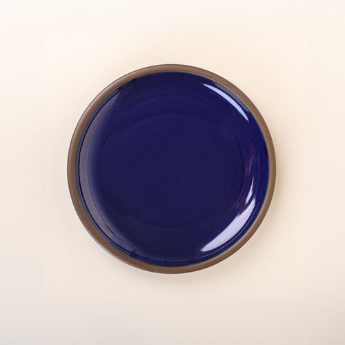A dinner sized ceramic plate in a glossy dark blue color featuring iron speckles and an unglazed rim