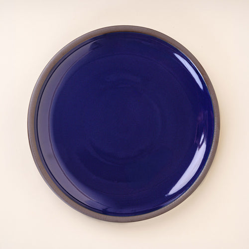 A large ceramic platter in a glossy dark blue color featuring iron speckles and an unglazed rim.