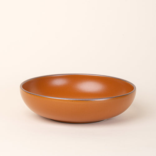 A large shallow serving ceramic bowl in an earthy orange color featuring iron speckles and an unglazed rim.