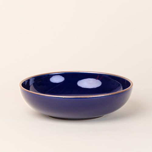 A large shallow serving ceramic bowl in a glossy dark blue color featuring iron speckles and an unglazed rim.