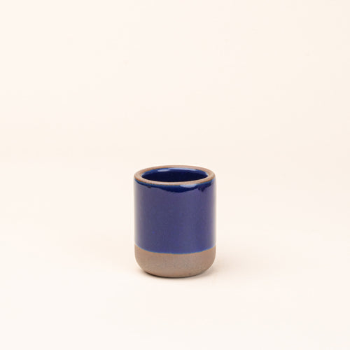 A small, short ceramic mug cup in a glossy dark blue color featuring iron speckles and unglazed rim and bottom base.