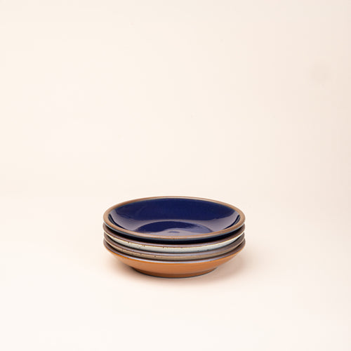 A stack of 4 small ceramic plates with a curved bowl edge in an earthy orange, glossy dark blue, white, and warm pale brown colors featuring iron speckles and an unglazed rim.