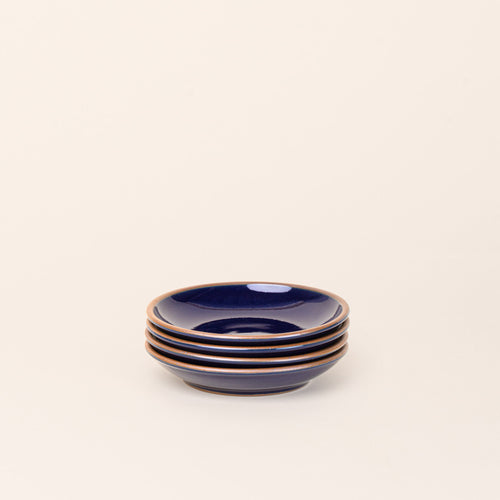 A stack of 4 small ceramic plates with a curved bowl edge in a glossy dark blue color featuring iron speckles and an unglazed rim.
