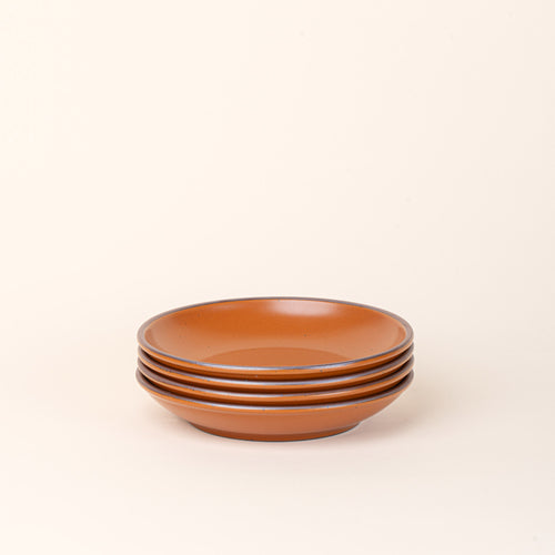A stack of 4 medium ceramic plates with a curved bowl edge in an earthy orange color featuring iron speckles and an unglazed rim.