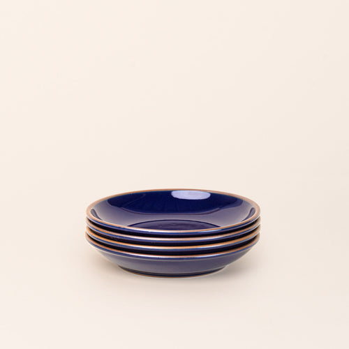 A stack of 4 medium ceramic plates with a curved bowl edge in a glossy dark blue color featuring iron speckles and an unglazed rim.