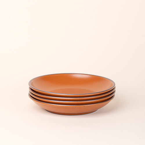 A stack of 4 large ceramic plates with a curved bowl edge in an earthy orange color featuring iron speckles and an unglazed rim.