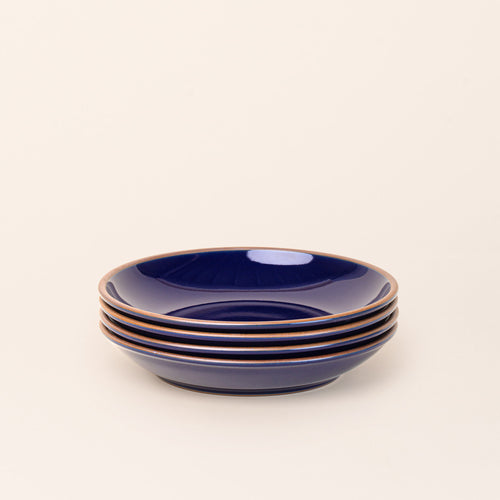 A stack of 4 large ceramic plates with a curved bowl edge in a glossy dark blue color featuring iron speckles and an unglazed rim.