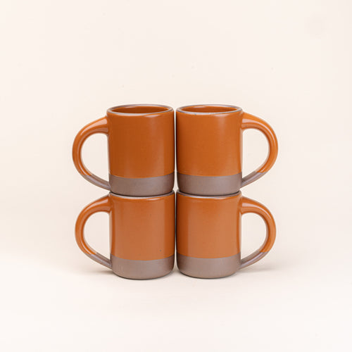 Four medium sized ceramic mugs with handles in an earthy orange color featuring iron speckles and unglazed rim and bottom base, stacked in sets of 2.