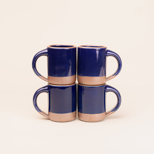 Four medium sized ceramic mugs with handles in a glossy dark blue color featuring iron speckles and unglazed rim and bottom base, stacked in sets of 2.