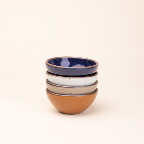 A stack of 4 small dessert sized rounded ceramic bowls in an earthy orange, glossy dark blue, white, and warm pale brown colors featuring iron speckles and an unglazed rim