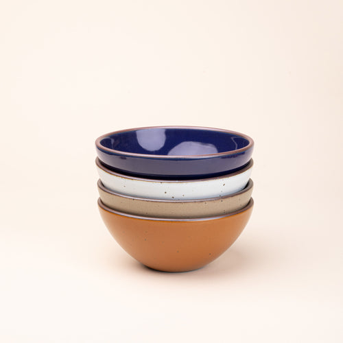 A stack of 4 medium sized rounded ceramic bowls in an earthy orange, glossy dark blue, white, and warm pale brown colors featuring iron speckles and an unglazed rim