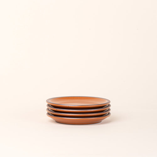 A stack of 4 dessert sized ceramic plates in an earthy orange color featuring an unglazed rim.