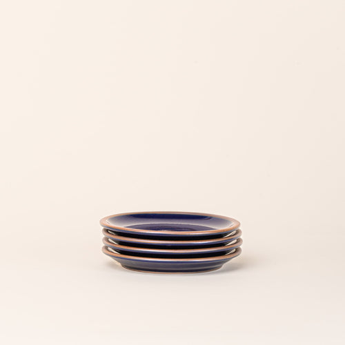 A stack of 4 dessert sized ceramic plates in a glossy dark blue color featuring an unglazed rim.