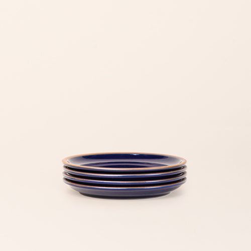 A stack of 4 medium sized ceramic plates in a glossy dark blue color featuring iron speckles and an unglazed rim.