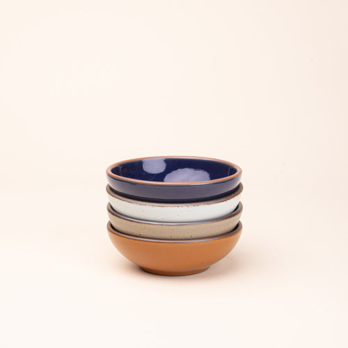 A stack of 4 small shallow ceramic bowls in an earthy orange, glossy dark blue, white, and warm pale brown colors featuring iron speckles and an unglazed rim