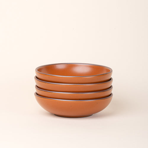 A stack of 4 dinner-sized shallow ceramic bowls in an earthy orange color featuring iron speckles and an unglazed rim